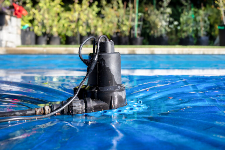 Automatic pool cover pump on top of blue wet cover in front of defocused garden foliage. Perspective view of black pool cover pump for keeping the water away from pool covers or spas. Selective focus.