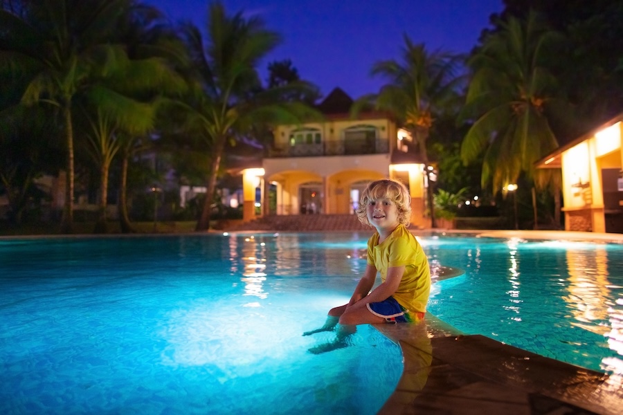 Kids play in outdoor swimming pool of tropical resort at night.