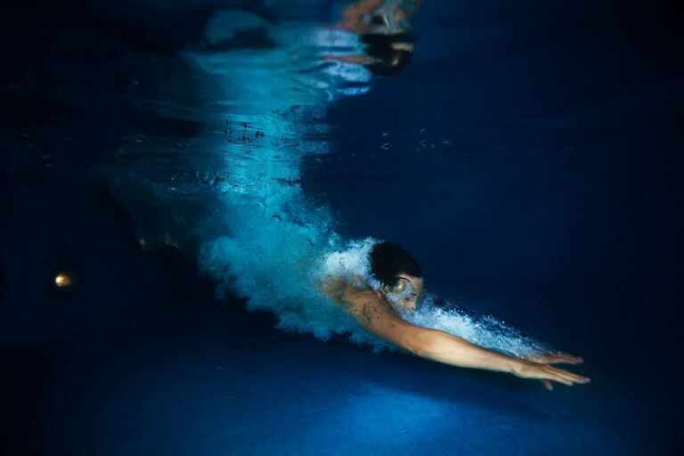 Man with white water splash swimming with straight arms under the clear dark blue water of swimming pool during night