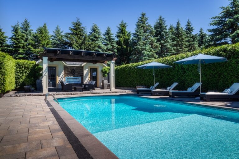 Luxury residential pool and cabana surrounded by tall hedges on a warm summer day.
