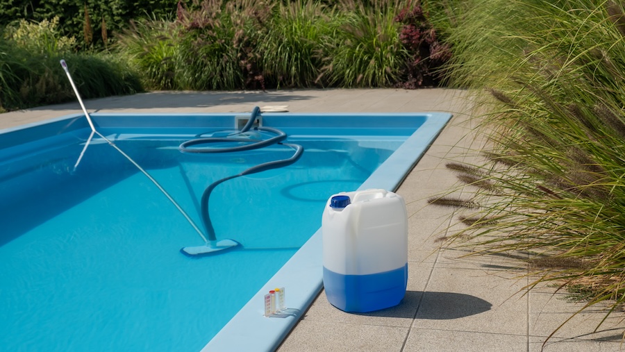 This image captures a pool cleaning hose and vacuum head submerged in a clear blue pool, with a large chemical container